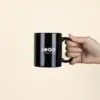 Mano sujetando una taza negra con asa personalizada con la palabra logo en blanco fotografiada sobre un fondo beige
