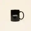 Taza negra con asa personalizada con la palabra logo en blanco fotografiada sobre un fondo beige
