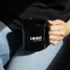 Chica en vaqueros agarrando una taza negra con asa personalizada con la palabra logo en blanco