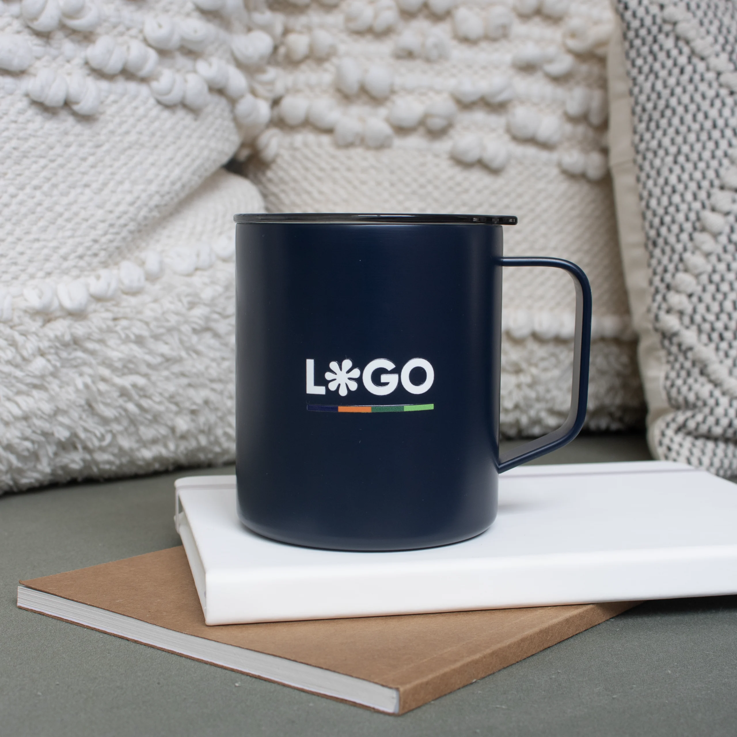 Vaso térmico con asa azul MACA Vaso térmico con asa azul con logo blanco apoyada en cuadernos