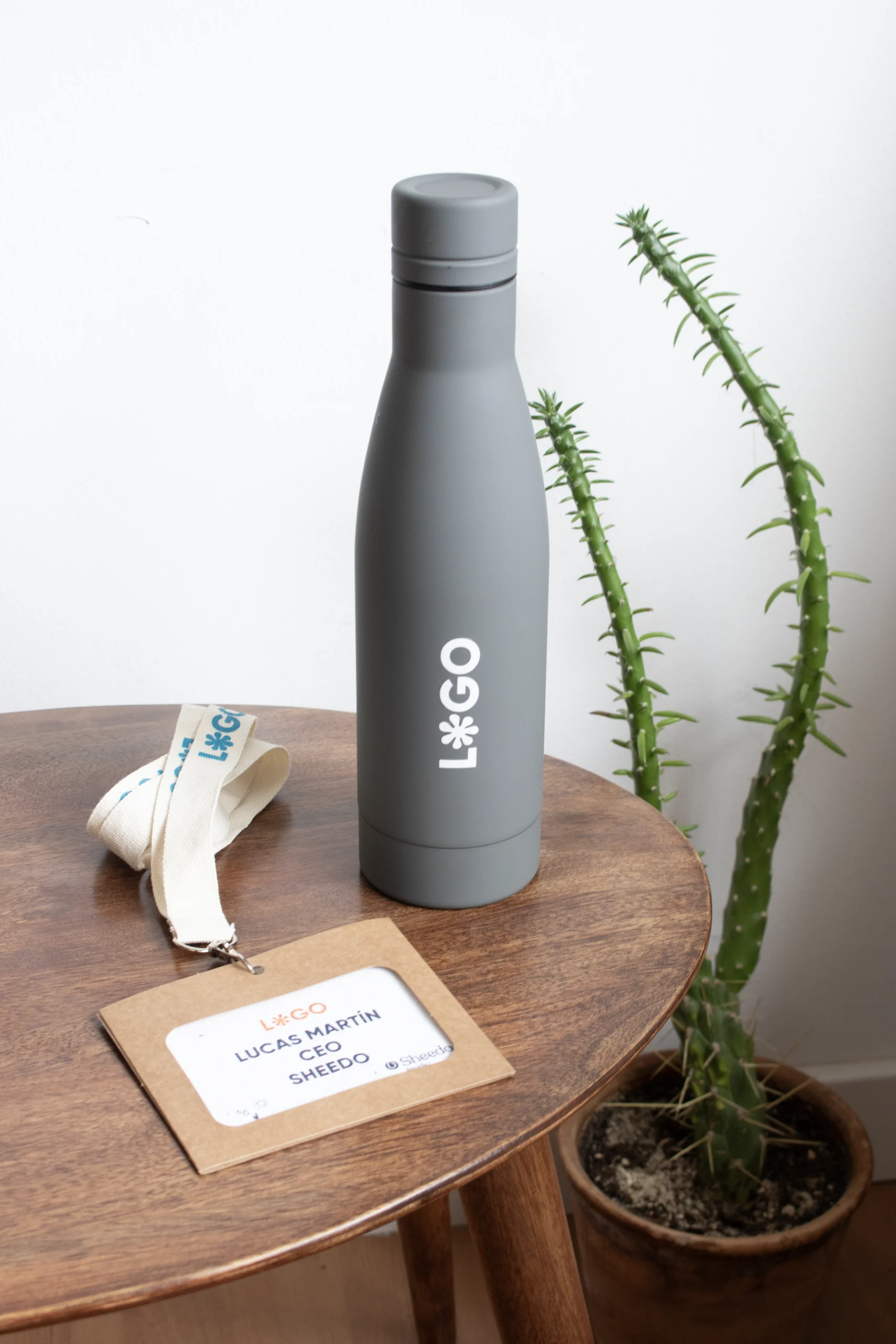 Gourde isotherme BALANCE Gris Botella gris de acero inoxidable personalizada con logo blanco junto a una acreditación con lanyard sobre una mesa de madera