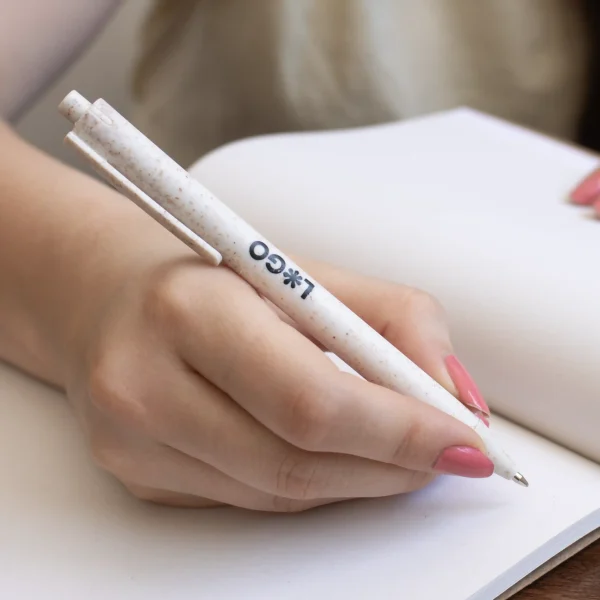 Mano escribiendo en cuaderno con bolígrafo de caña de trigo blanco personalizado con logo