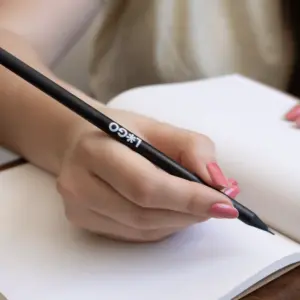 Mano escribiendo en cuaderno con lápiz de madera negro personalizado con logo blanco