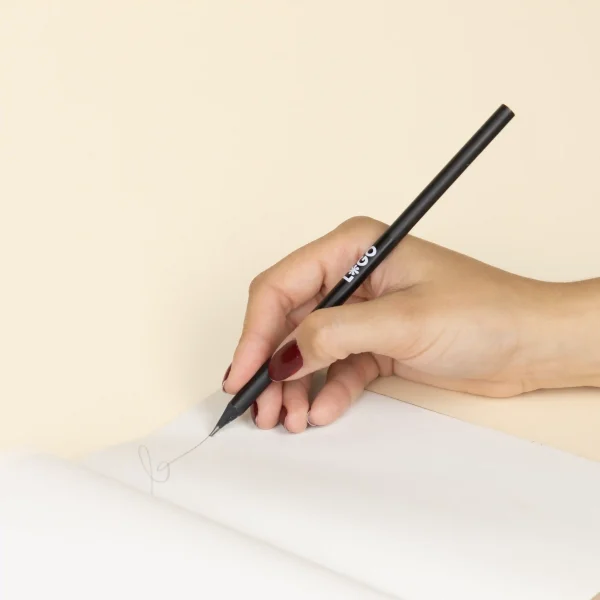 Mano escribiendo en cuaderno con lápiz de madera negro personalizado con logo blanco fondo beige
