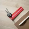 Llaves con llavero de tela rojo bordado con hilo blanco sobre una mesa de madera junto a un cuaderno y un bolígrafo