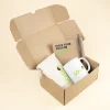 Caja de cartón abierta con un neceser beige, una taza blanca, un cuaderno kraft y un bolígrafo marrón personalizados con logo