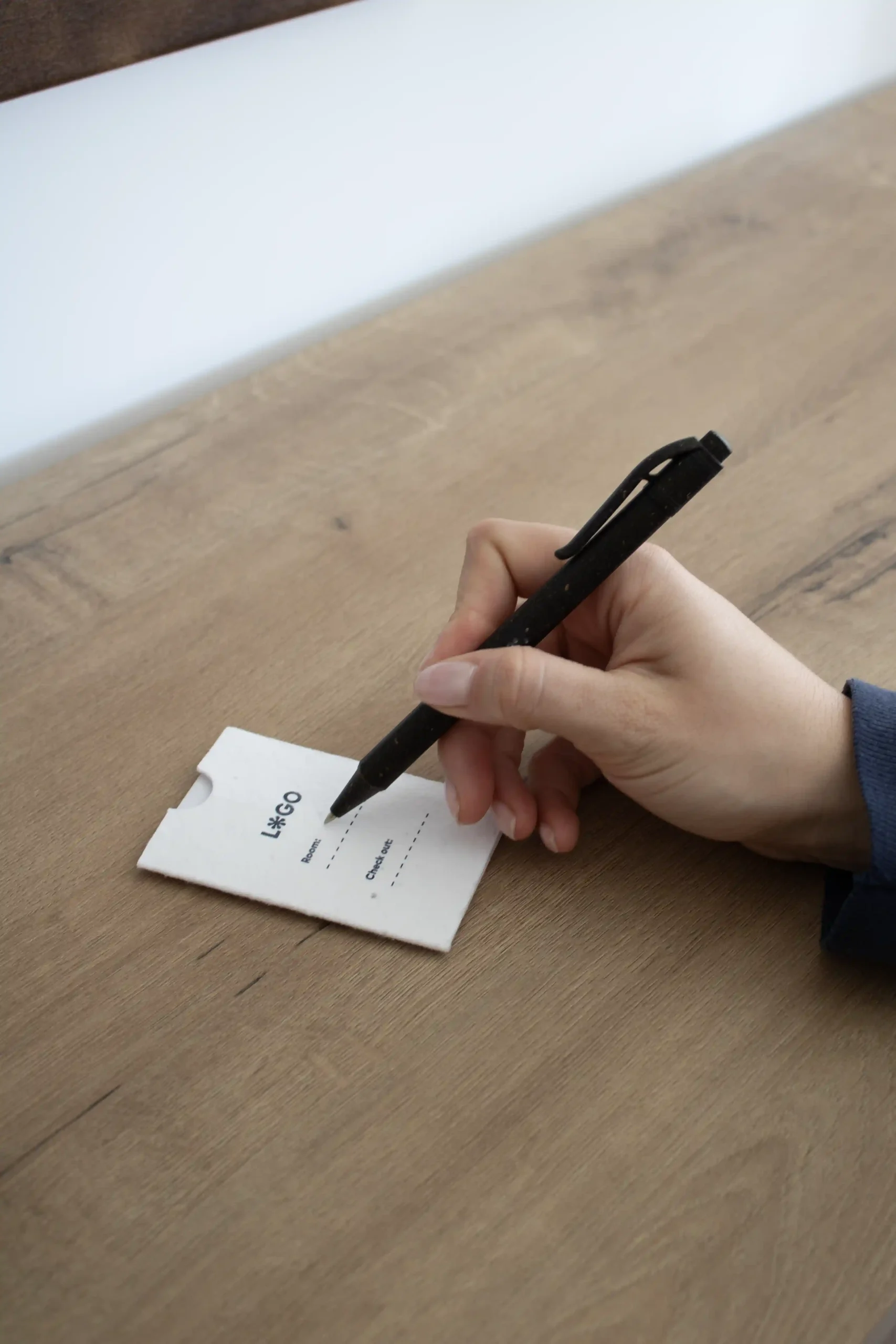 Porta llaves hotel sobre SAK + Bolígrafo ERBA mano escribiendo en una mesa de madera con un bolígrafo en un porta llaves de hotel blanco personalizado con la palabra logo en azul