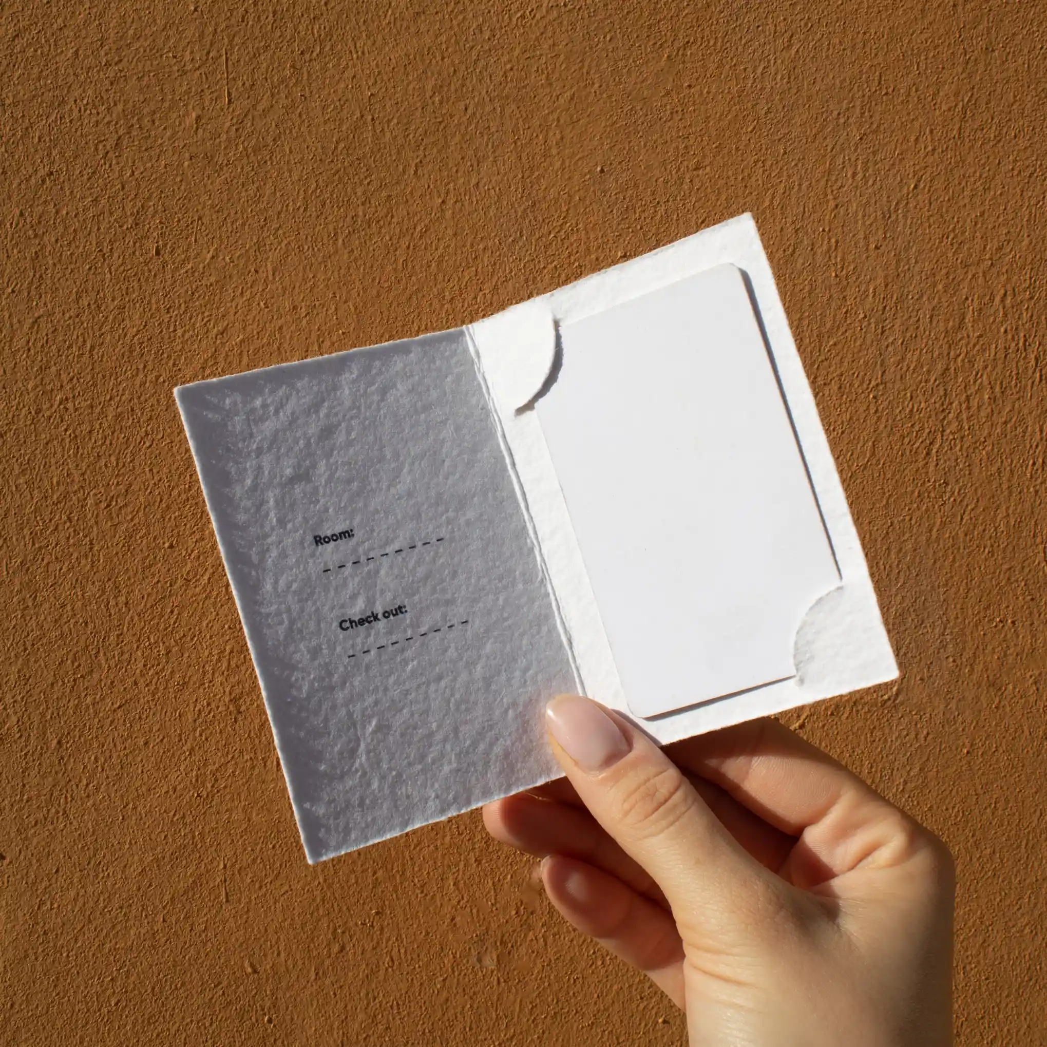 Mano mostrando un porta llaves de hotel díptico en papel ecológico blanco abierto con una tarjeta blanca dentro