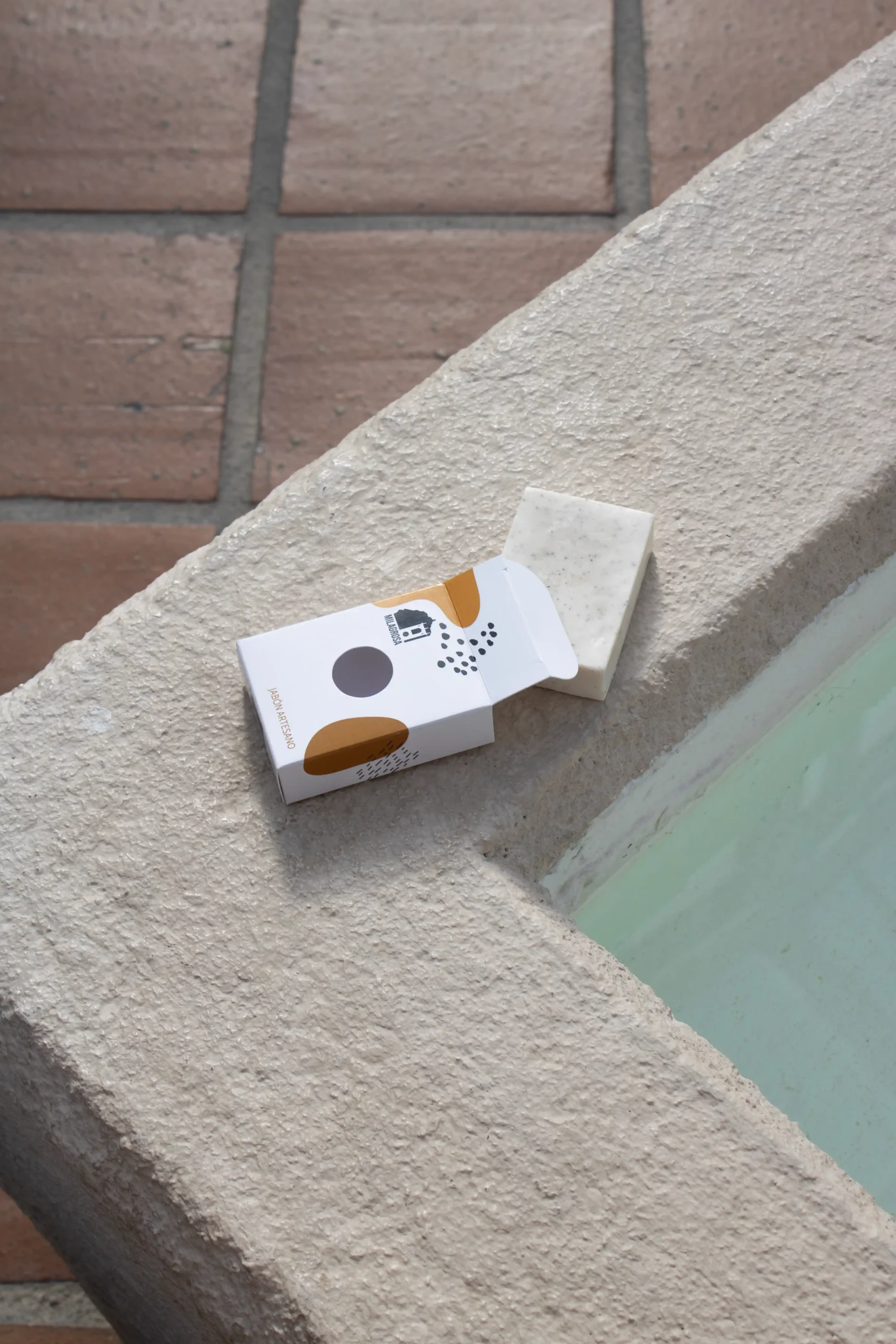 Jabón artesanal dentro de una caja blanca personalizada para el Hotel La Milagrosa, apoyado en el borde de una piscina