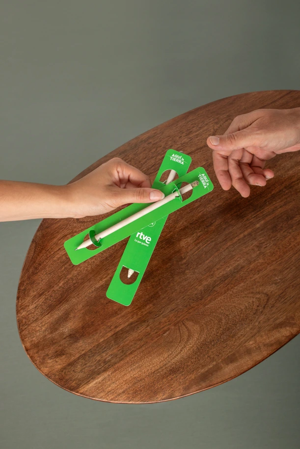 Manos sujetando dos lápices plantables personalizados en un packaging verde sobre una mesa de madera y un fondo de color verde