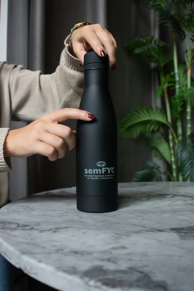 Manos de mujer mostrando una botella negra personalizada apoyada en una mesa de mármol con una planta al fondo