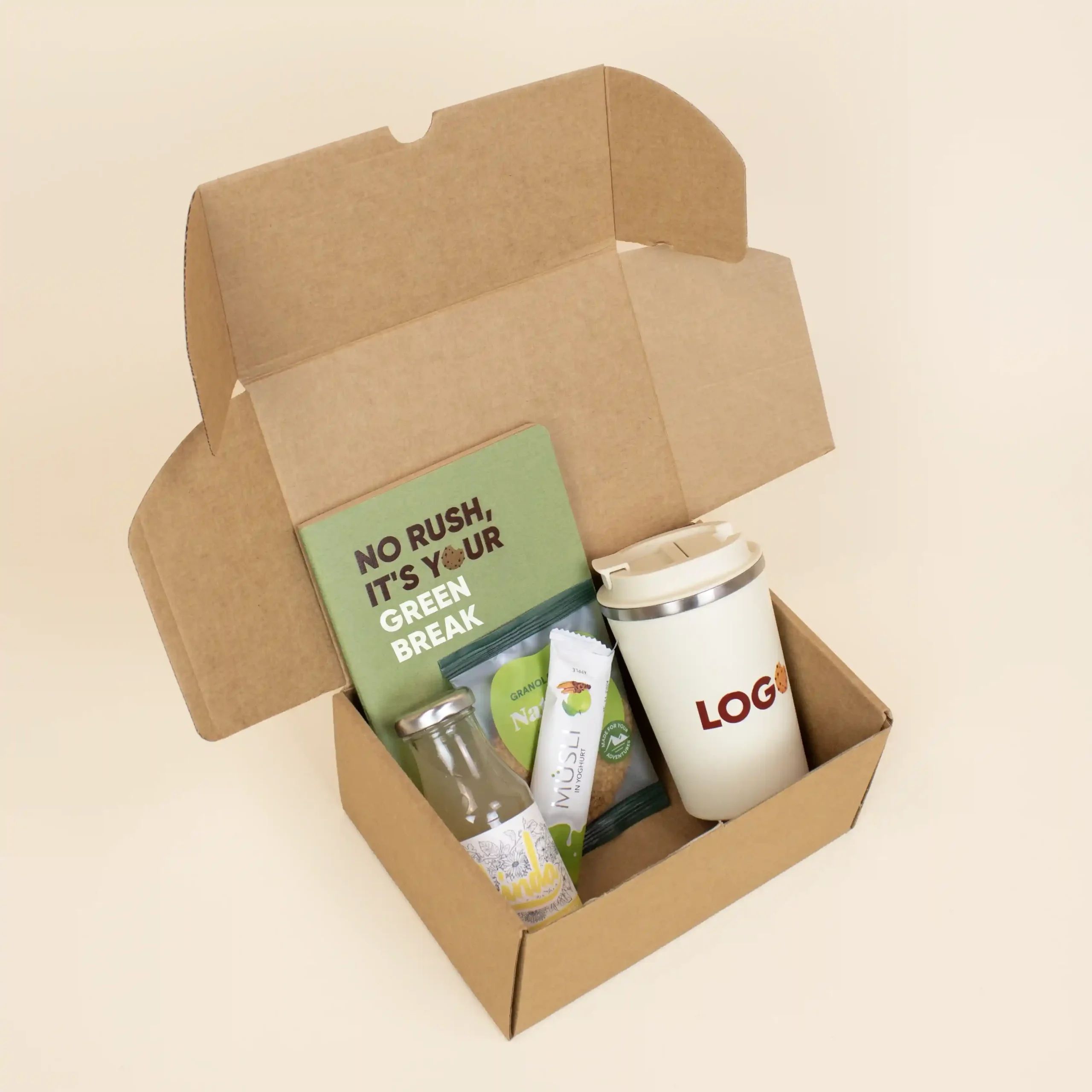 Caja de cartón abierta con una taza take away, un cuaderno verde, una limonada, una barrita de muesli y una galleta