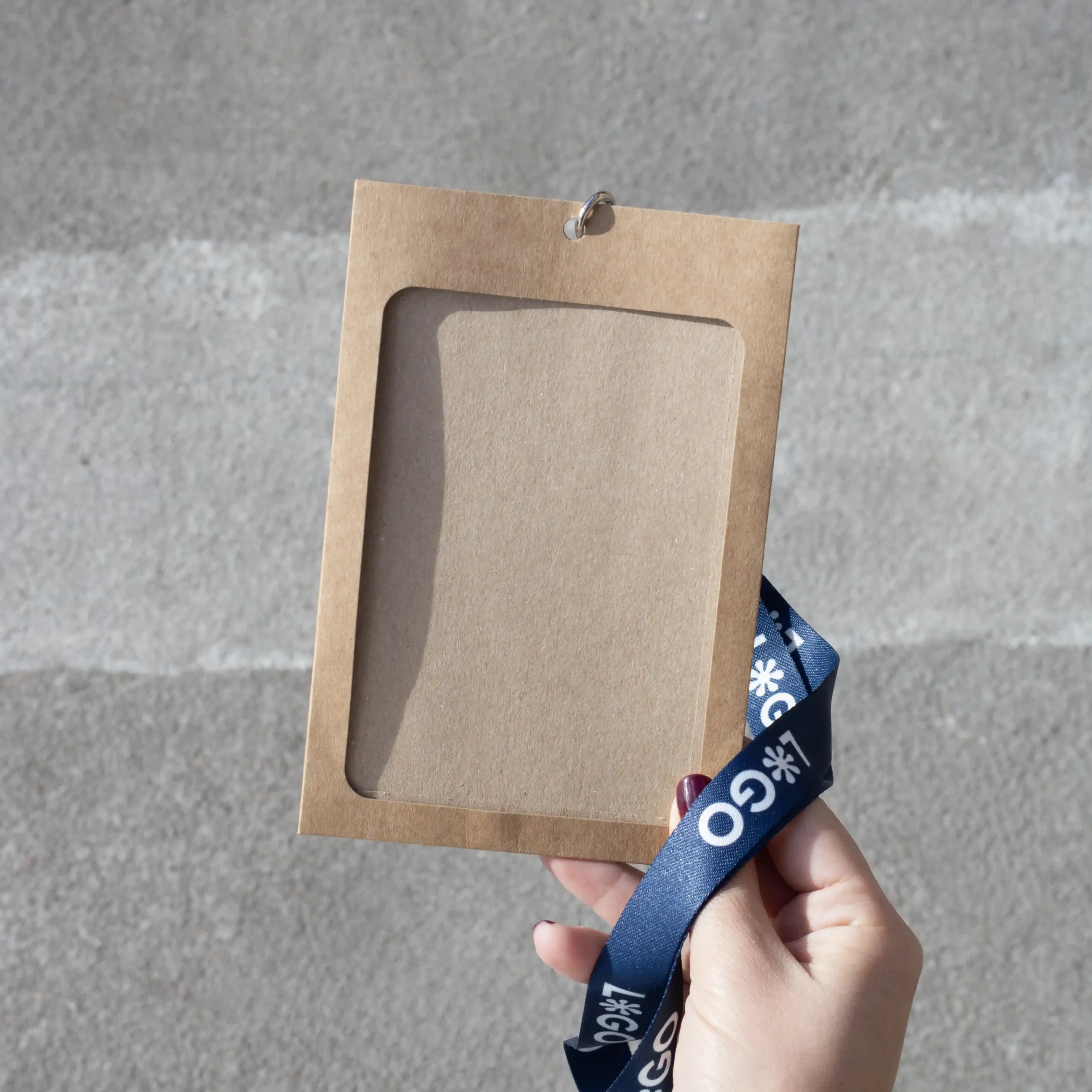 Kit B: Funda KASE Kraft 0/0 + Lanyard COLLIN Mano sujetando una funda de acreditación kraft con lanyard de rPET azul personalizado con el texto 'logo' en blanco