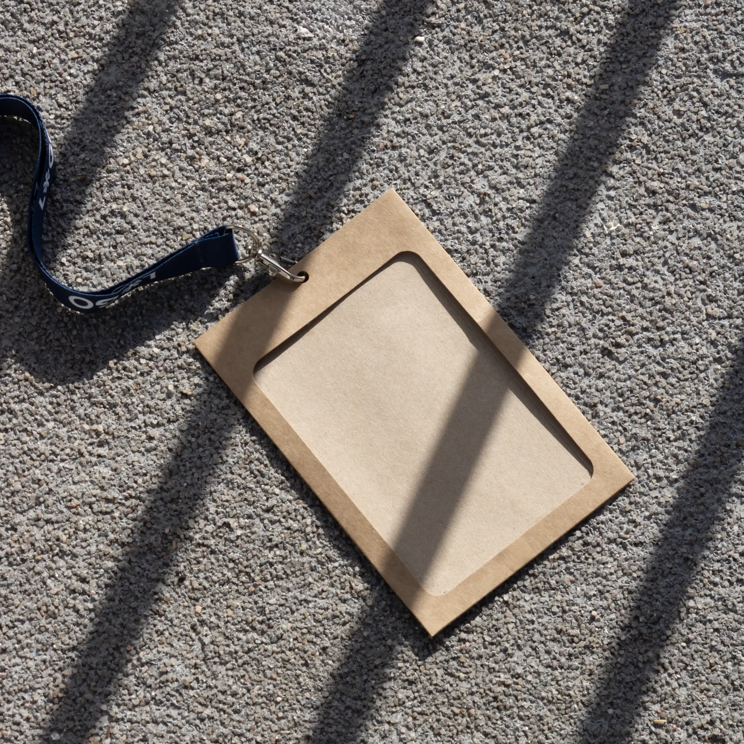 Kit B: Funda KASE Kraft 0/0 + Lanyard COLLIN Funda de acreditación kraft con lanyard de rPET azul apoyado en el suelo de la calle