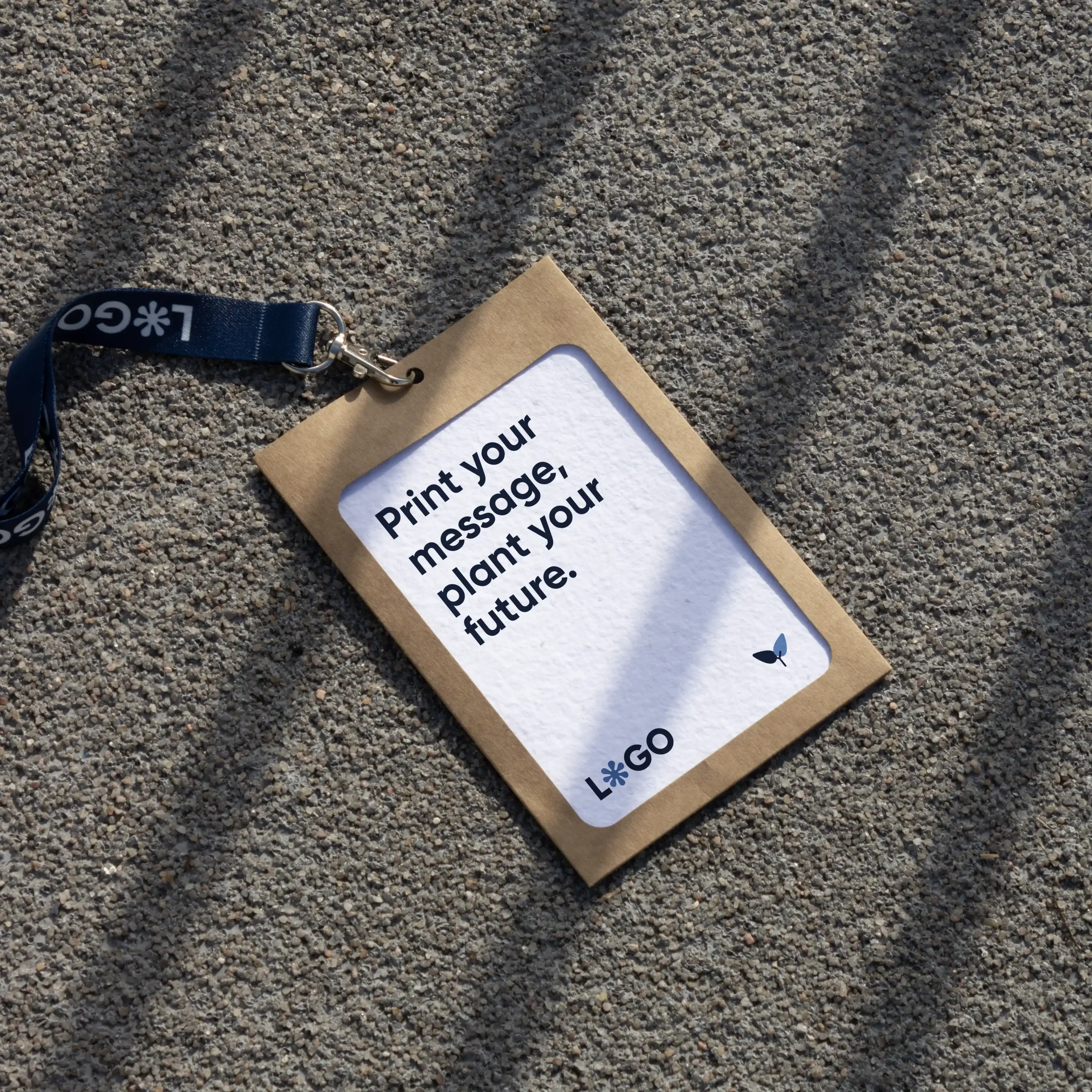 Kit C: Acreditación plantable, Funda KASE 0/0 y Lanyard COLLIN Acreditación personalizada con funda kraft y lanyard de rPET azul personalizado con el texto 'logo' en blanco apoyada en el suelo de la calle