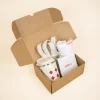 Caja de cartón abierta con una taza take away, un flyer, una camiseta blanca y una tote bag de tela