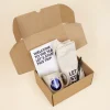 Caja de cartón abierta con un cuaderno blanco plantable, una taza de cerámica beige, un bolígrafo ecológico marrón, y una tote bag de tela personalizados