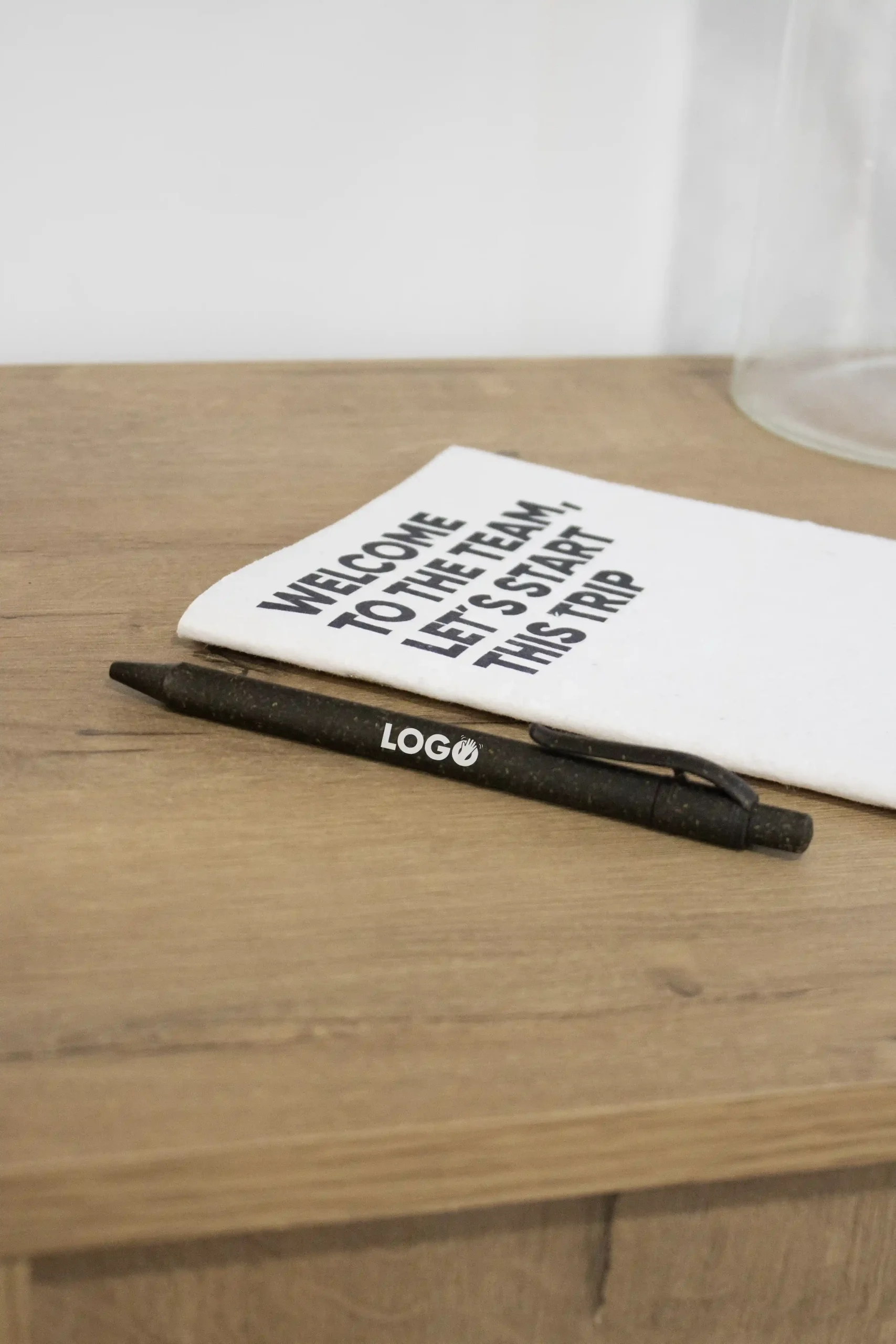 Pack 'Welcome to the team': ERBA Bolígrafo ecológico de hierba personalizado con la palabra logo en blanco junto a un cuaderno plantable blanco personalizado sobre una superficie de madera