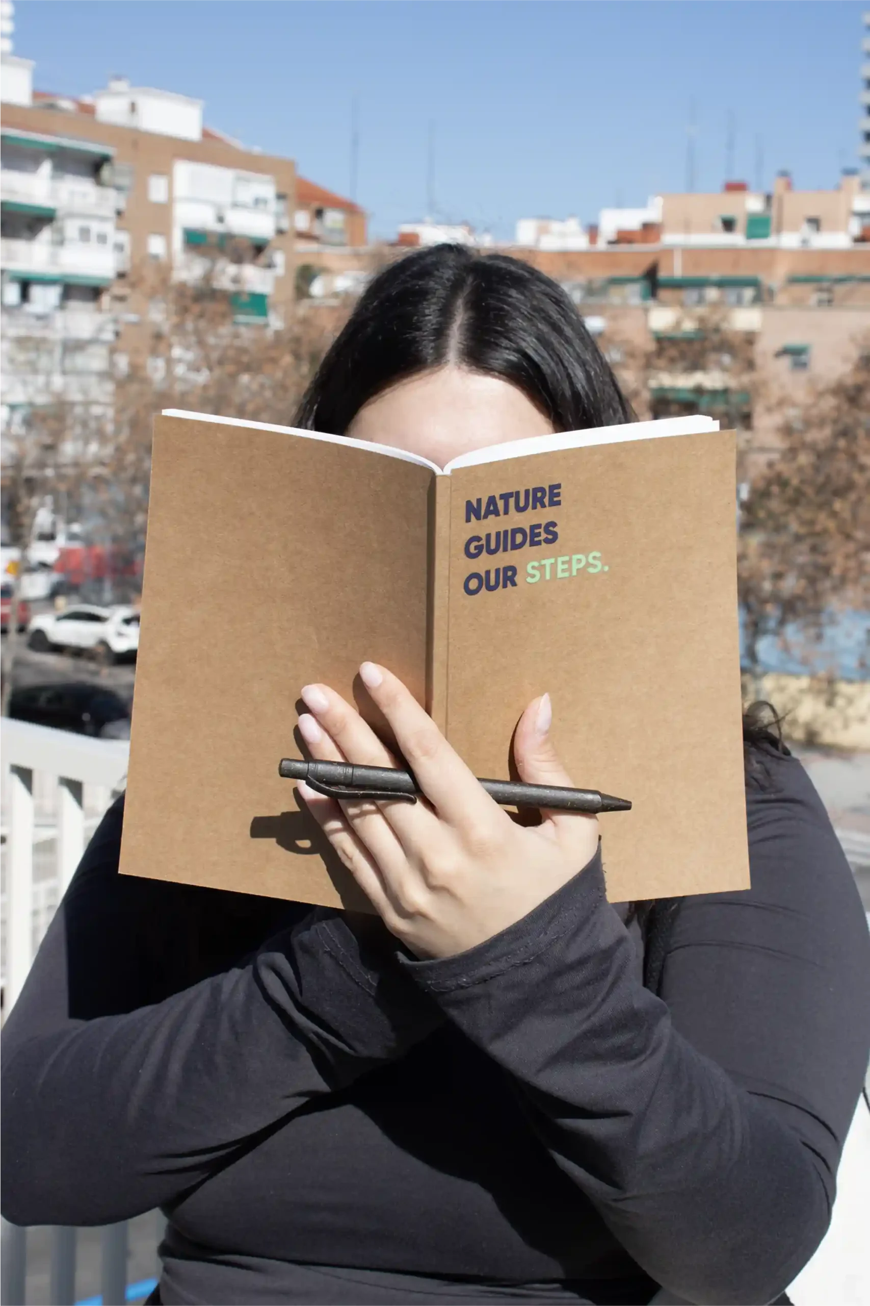 Carnet écologique KLAAS + Stylo ERBA Chica sujetando un cuaderno ecológico kraft personalizado con el texto 'Nature guides our steps' y un bolígrafo marrón plantable