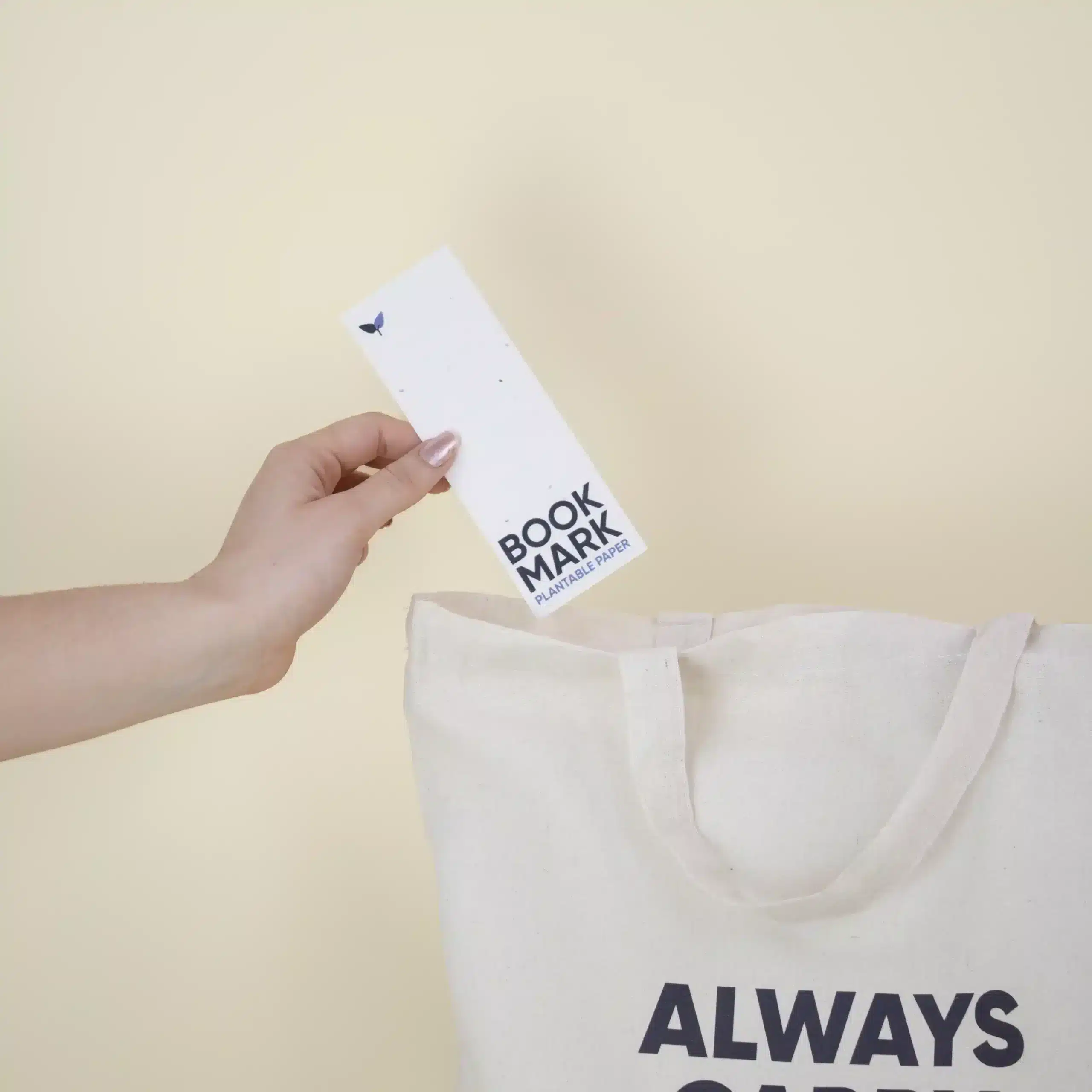 KIT CARRY A BOOK: Tote bag + Marcapáginas Mano metiendo un marcapáginas de papel plantable personalizado en una bolsa de algodón beige