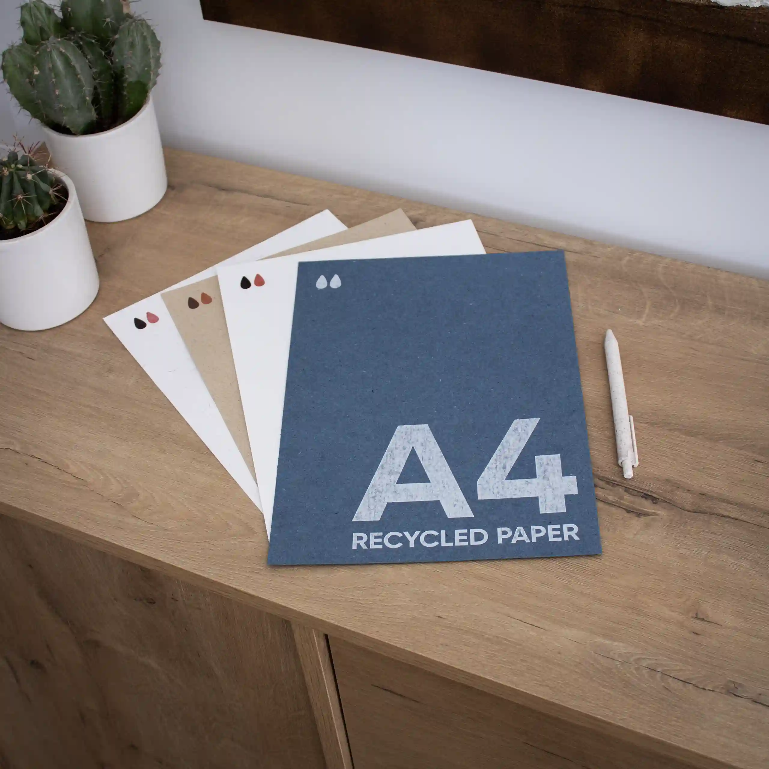 Cuatro tarjetas A4 impresas en papel ecológico junto a un bolígrafo sobre una superficie de madera