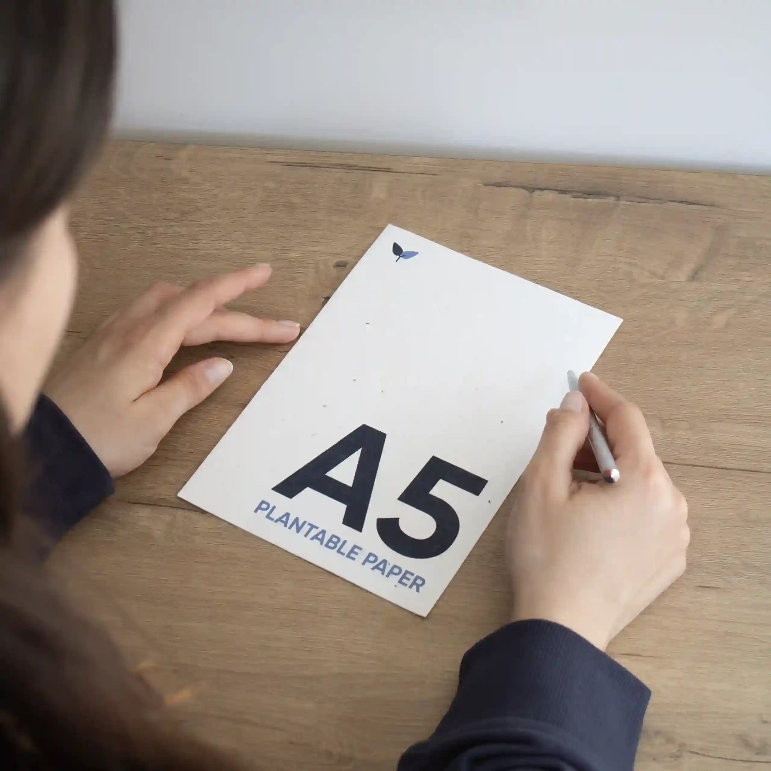 Tarjeta A5 plantable Chica de espaldas sentada en una mesa de madera escribiendo con un bolígrafo en una tarjeta A5 impresa en papel plantable