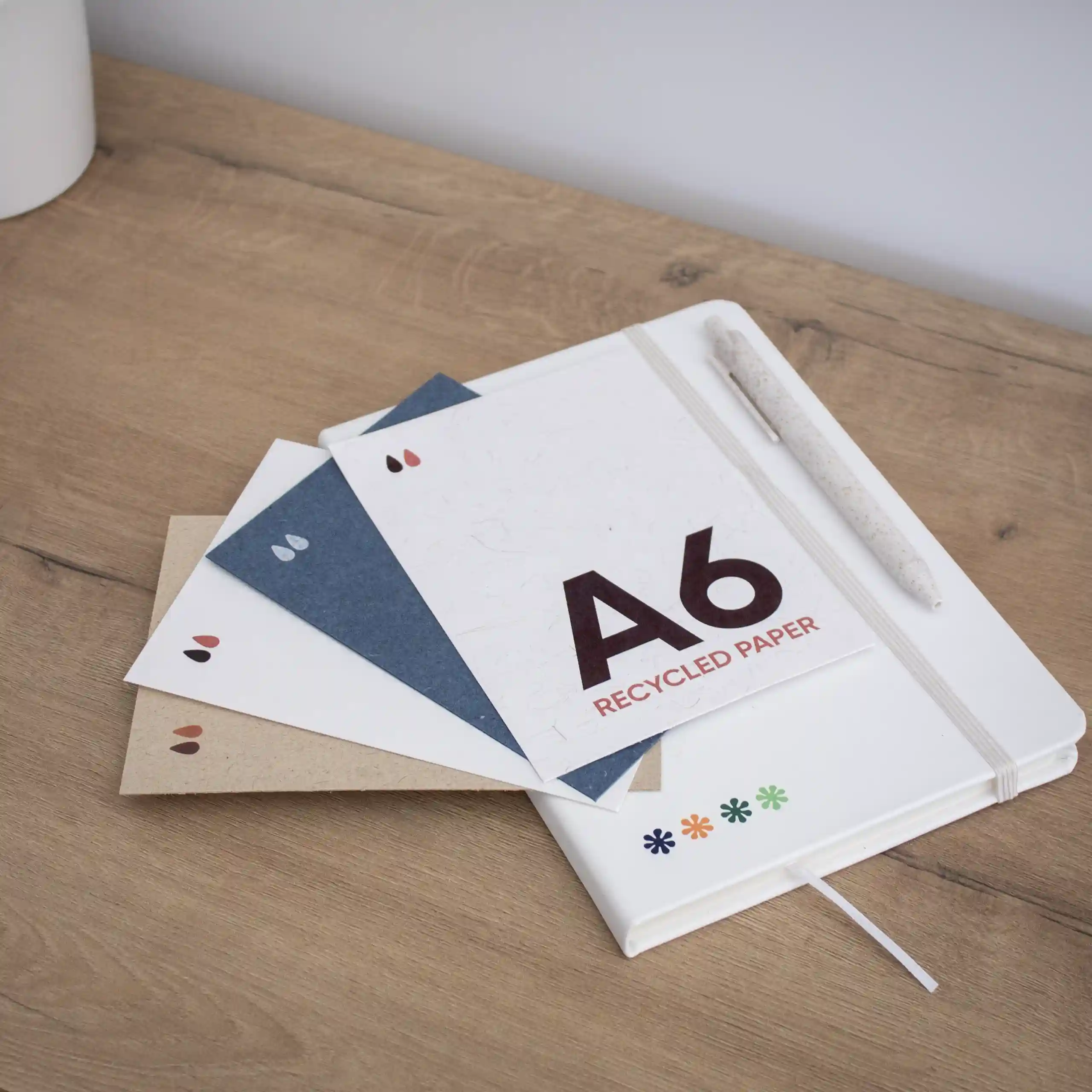 Tarjeta papel ecológico A6 cuatro tarjetas A6 impresas en papeles ecológicos junto a un cuaderno blanco y un bolígrafo, sobre una superficie de madera