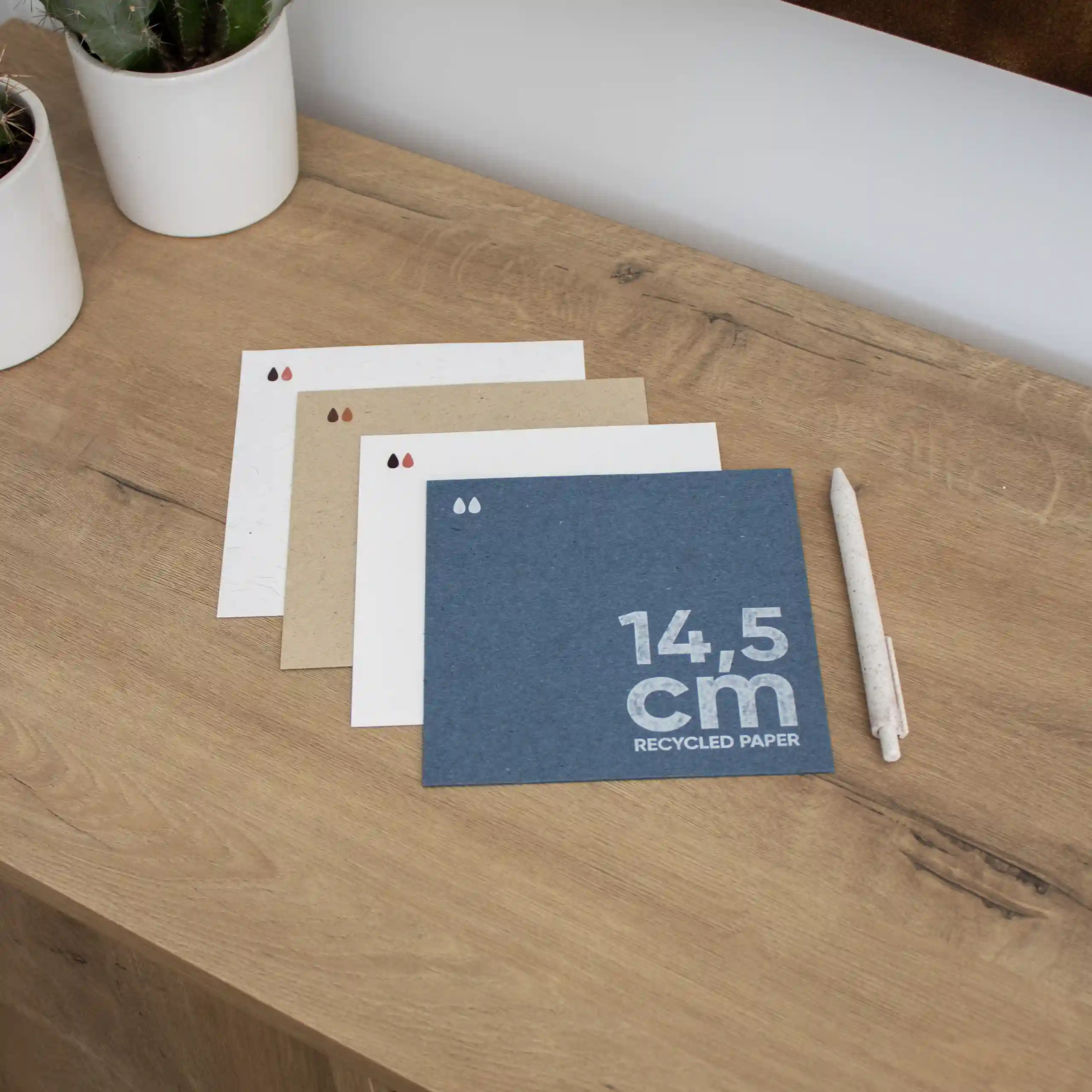 Cuatro tarjetas cuadradas impresas en papel ecológico junto a un bolígrafo sobre una superficie de madera
