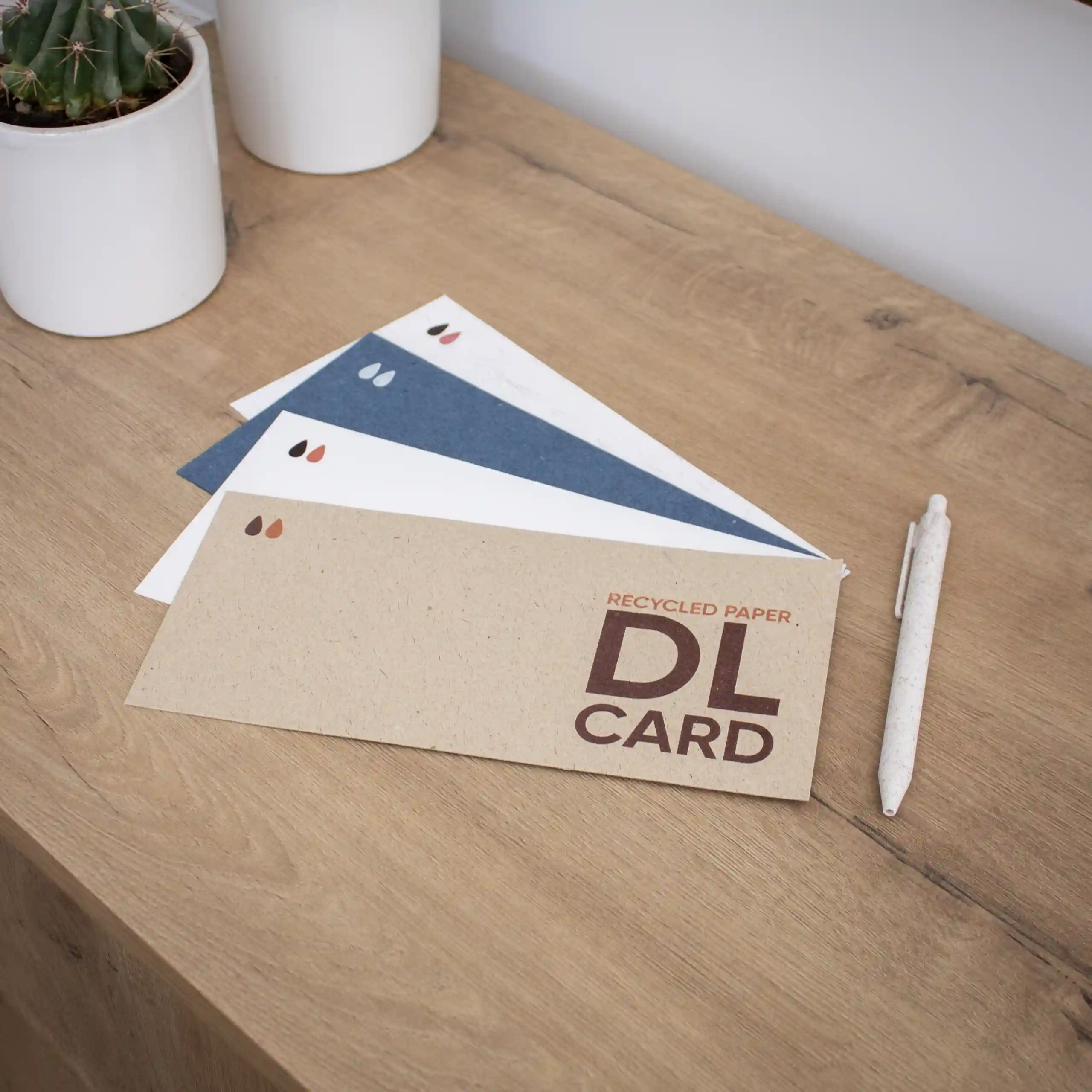 Cuatro tarjetas DL impresas en papel ecológico junto a un bolígrafo sobre una superficie de madera