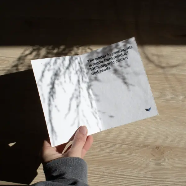 Mano sujetando un díptico A6 abierto impreso en papel plantable sobre una superficie de madera con sombras