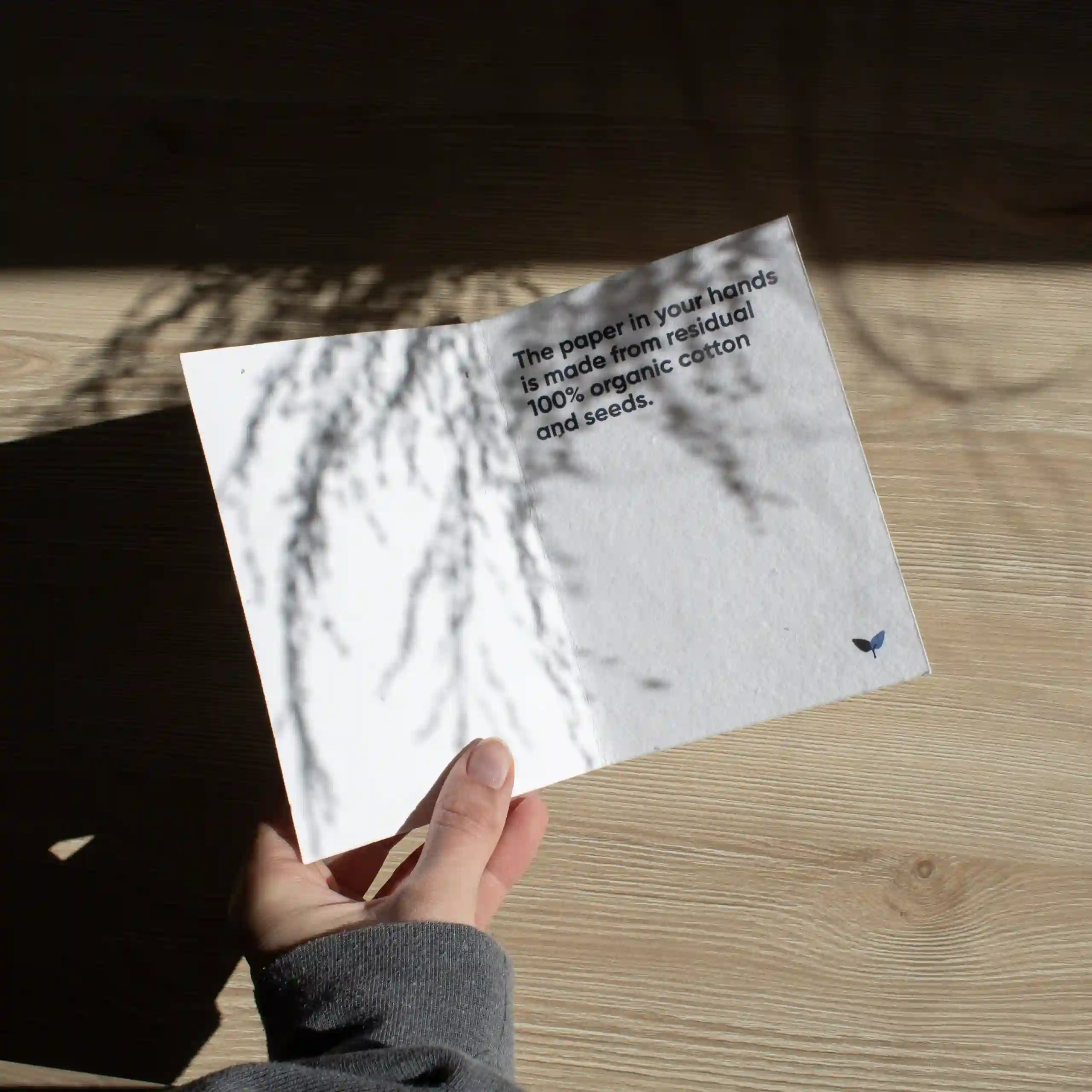 Carte pliante A6 plantable Mano sujetando un díptico A6 abierto impreso en papel plantable sobre una superficie de madera con sombras