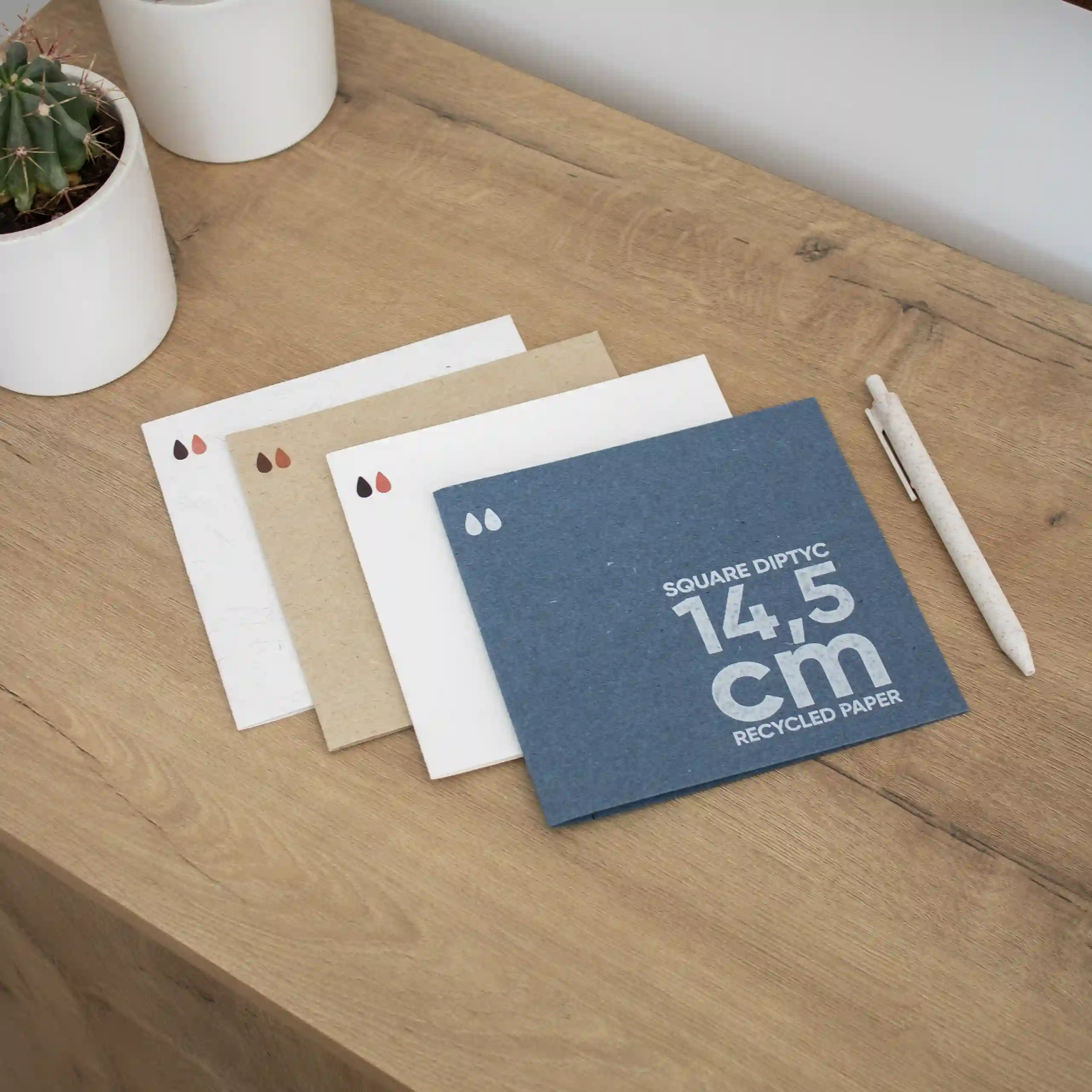 Díptico de papel ecológico cuadrado Cuatro dípticos cuadrados impresos en papel ecológico junto a un bolígrafo sobre un superficie de madera