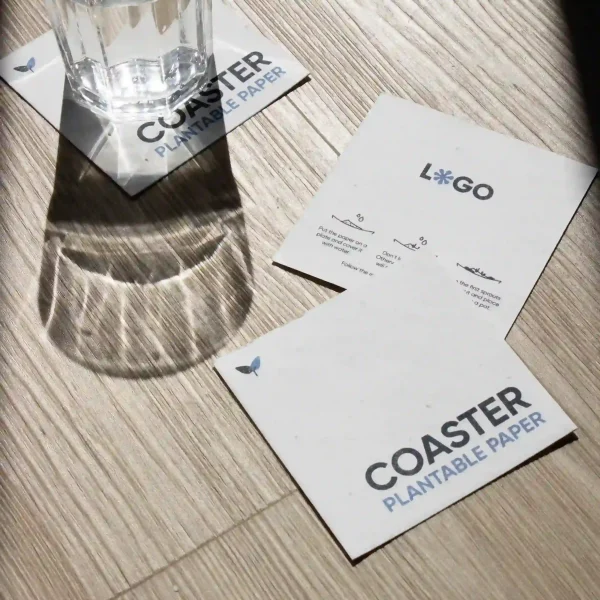 Tres posavasos de papel plantable personalizados y un vaso de cristal sobre una superficie de madera al sol
