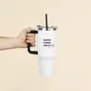 Mano sujetando un vaso térmico blanco personalizado de acero inoxidable con pajita sobre un fondo beige
