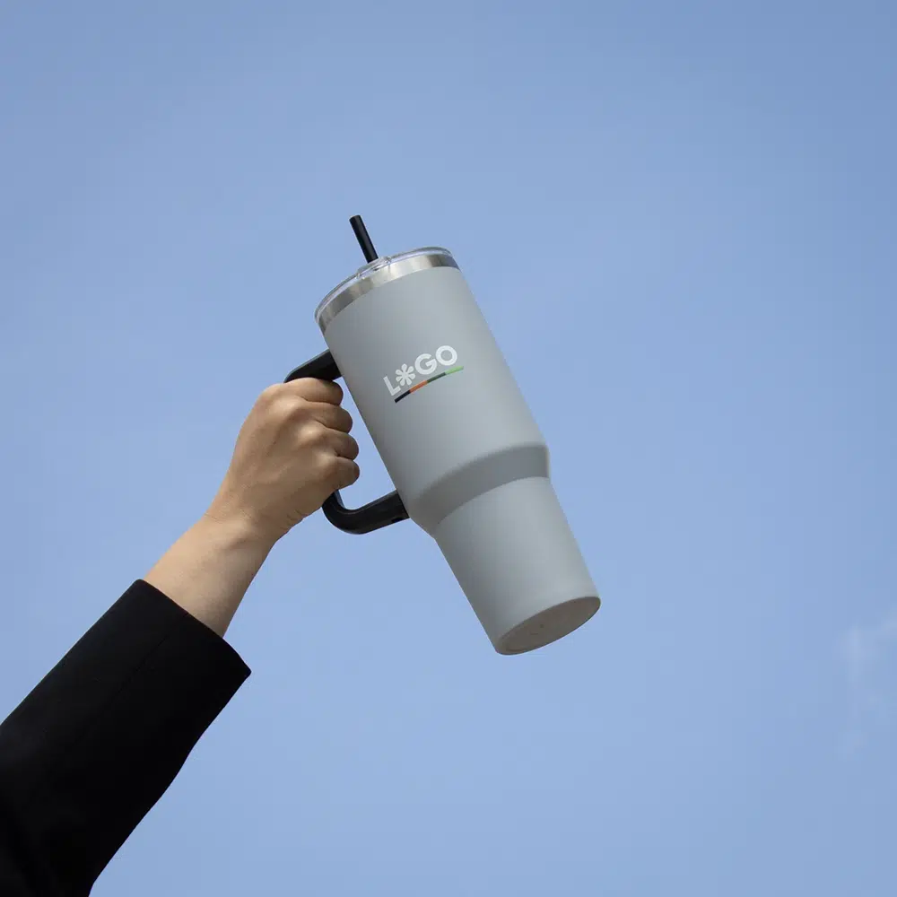 Travel tumbler EVEREST Mano alzando un vaso térmico gris de acero inoxidable con pajita con el cielo de fondo