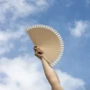 Mano sujetando un abanico de madera y algodón con el cielo de fondo