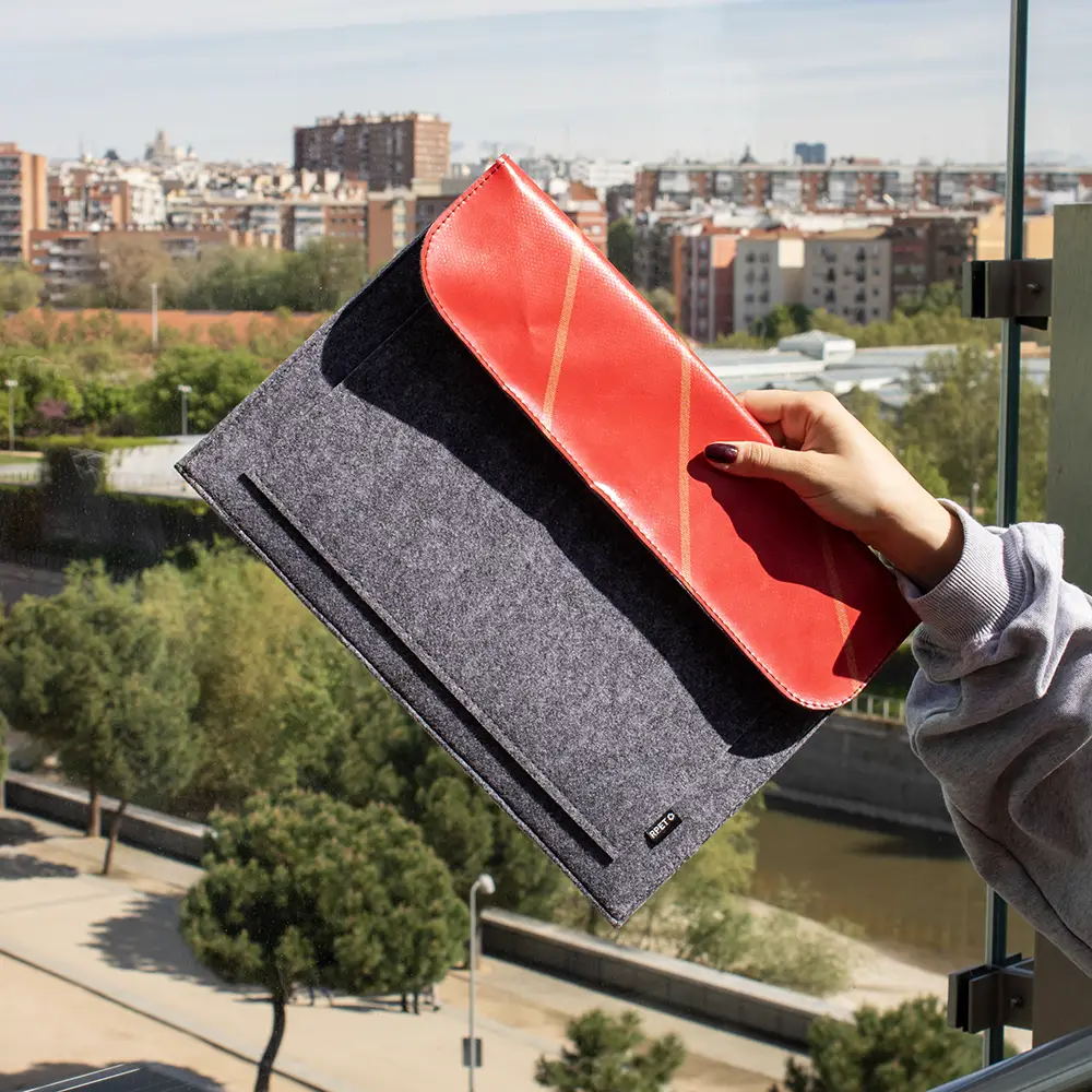Mano mostrando una funda de portátil personalizada hecha con fieltro y lona roja reciclada, con la ciudad de fondo