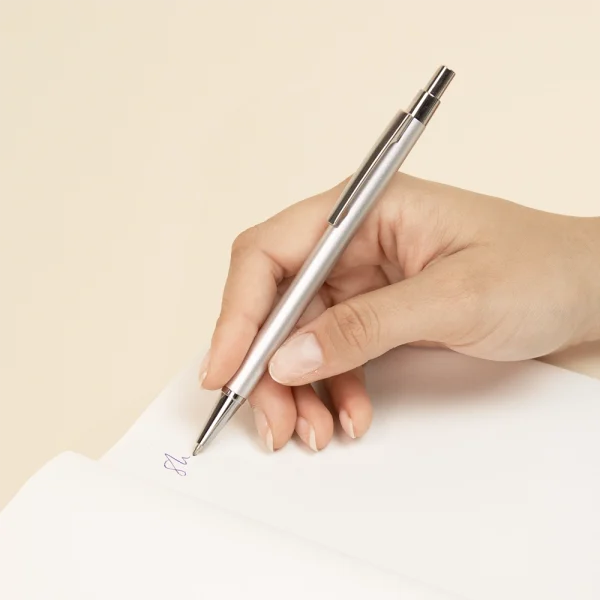 Mano escribiendo sobre un cuaderno con un bolígrafo plateado de aluminio reciclado