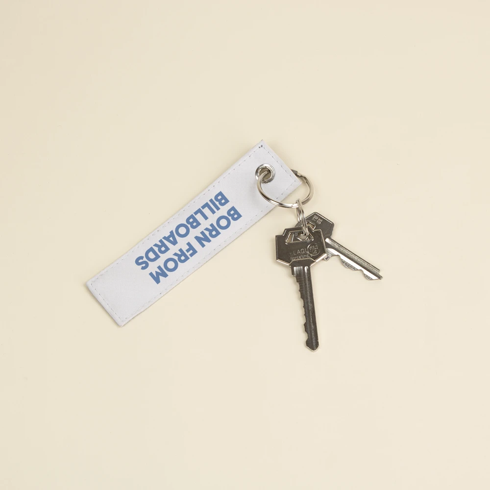 Llavero sostenible de lona LANIN Llavero hecho con lona reciclada blanca personalizado con el texto impreso en azul: "Born from billboards" con dos llaves enganchadas de su anilla metálica