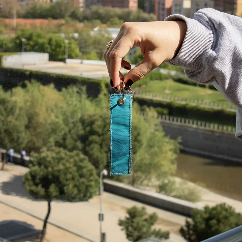 Llavero sostenible de lona LANIN Mano mostrando un llavero azul hecho con lona reciclada, con la ciudad de fondo