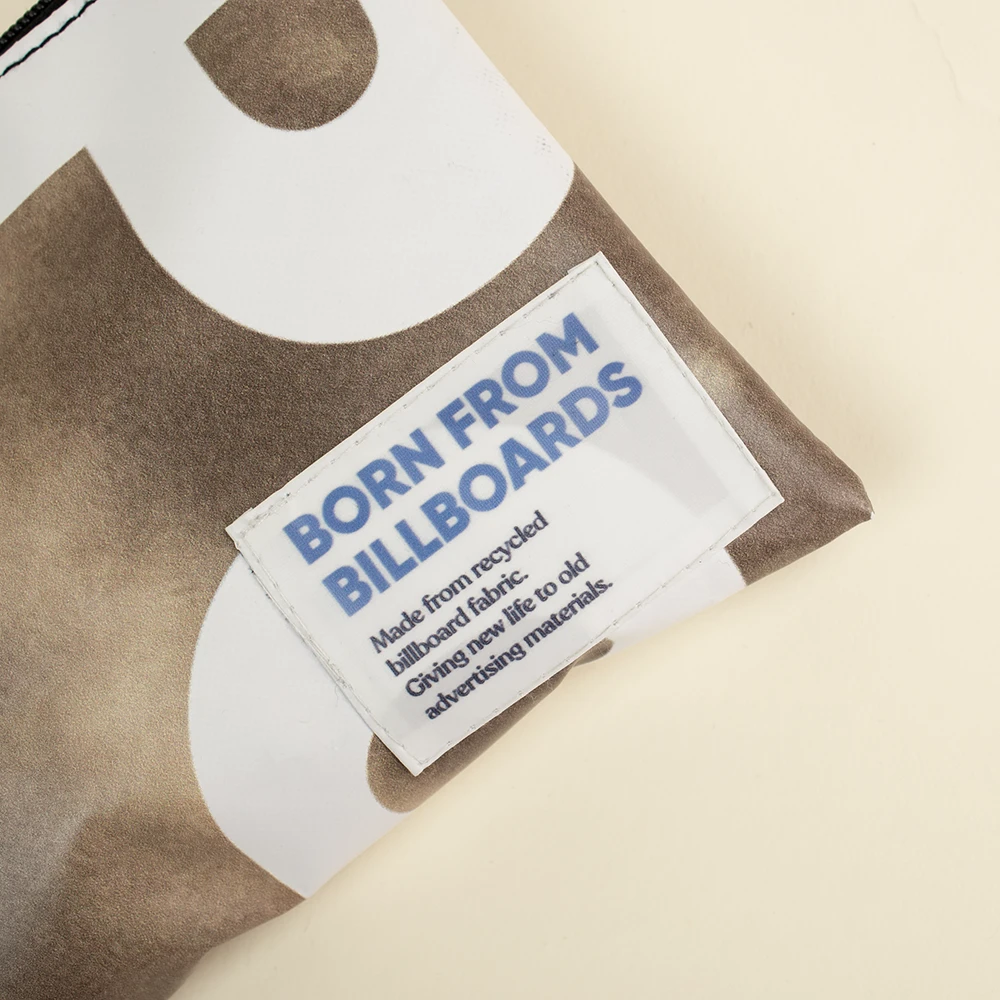 Etiqueta impresa con el texto "Born from Billboards" cosida un neceser de lona reciclada de color marrón y blanco