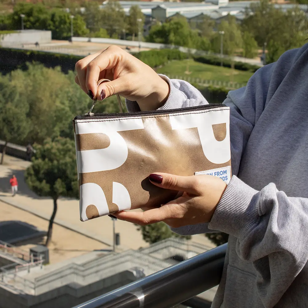 Chica abriendo un neceser de lona reciclada de color marrón y blanco, con la ciudad de fondo