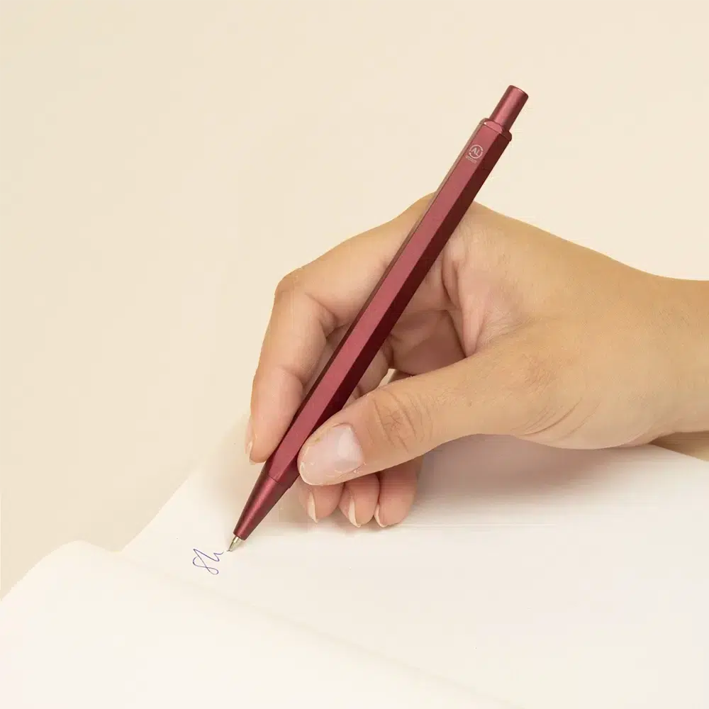 Bolígrafo hexagonal de aluminio SIXTO Mano escribiendo sobre un cuaderno con bolígrafo de aluminio rojo sobre un fondo beige