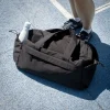 Bolsa de deporte negra de poliéster reciclado junto a unos pies con una botella blanca en uno de sus bolsillos