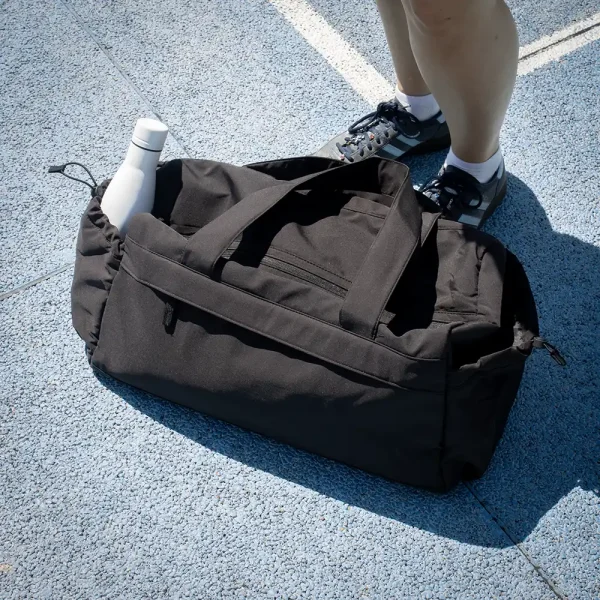 Bolsa de deporte negra de poliéster reciclado junto a unos pies con una botella blanca en uno de sus bolsillos