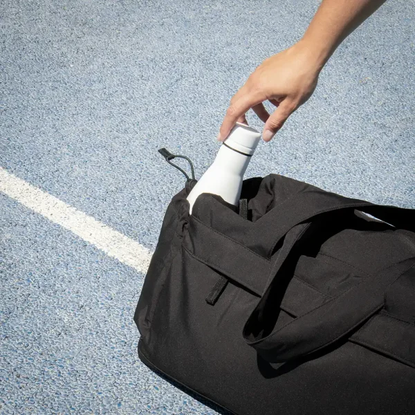 Mano cogiendo una botella blanca de una bolsa de deporte negra de poliéster reciclado.