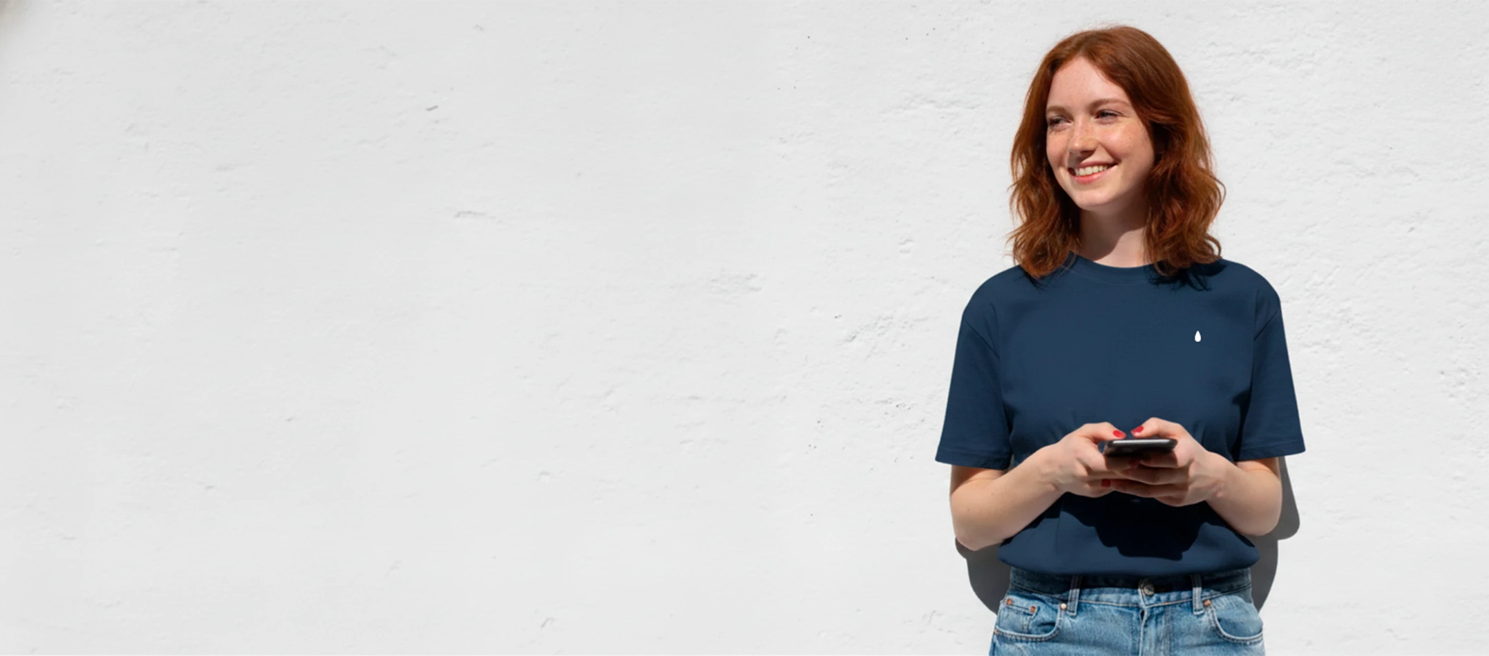 Chica pelirroja sonriendo con una camiseta azul personalizada y con un móvil en las manos, apoyada en una pared blanca