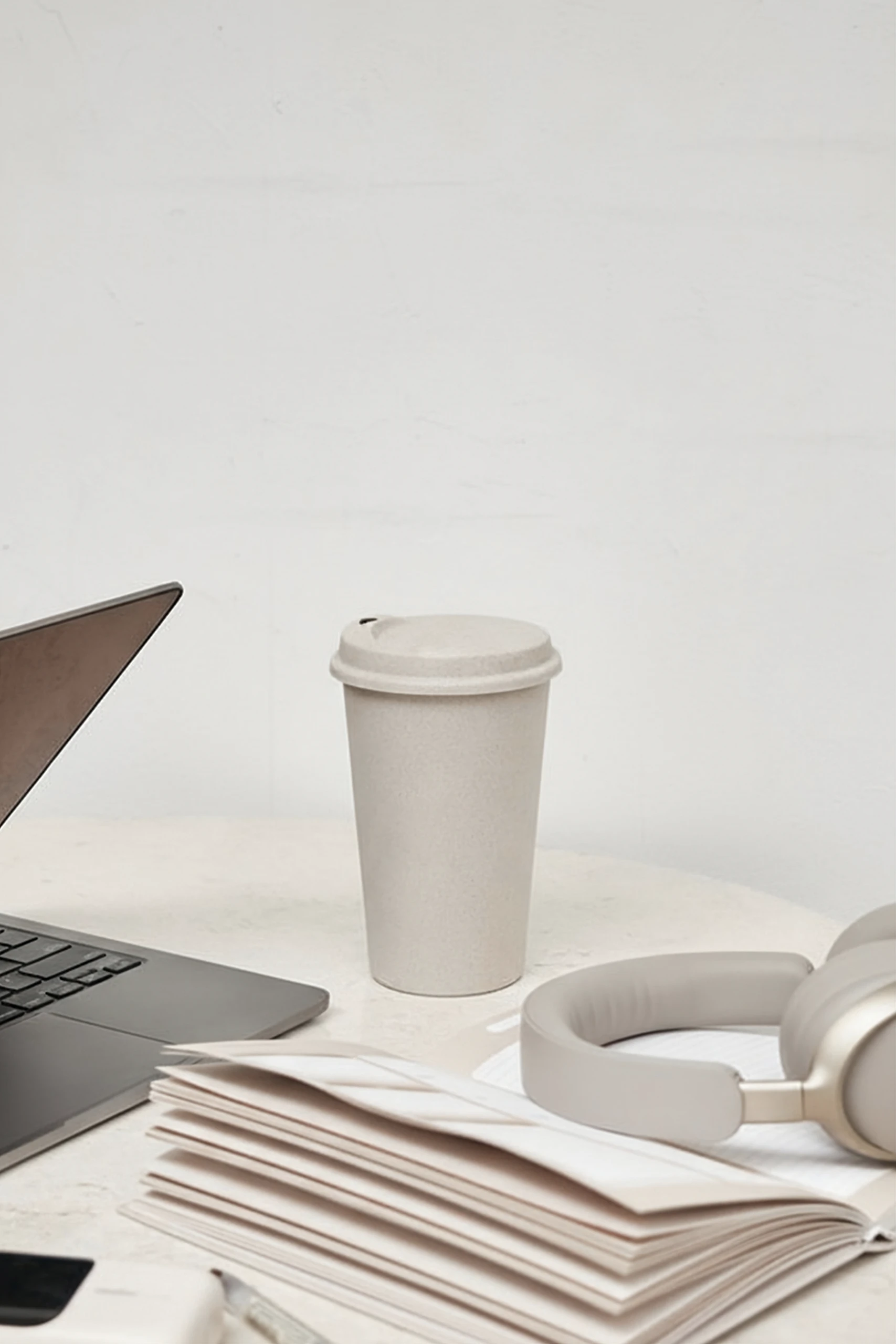 Vaso reutilizable take away con tapa apoyado sobre una mesa de trabajo junto a un portátil, un cuaderno y unos cascos