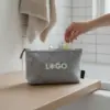 Neceser de viaje personalizado ARLO Mano metiendo un cosmético dentro de un neceser de viaje de color gris personalizado con la palabra logo en blanco sobre una superficie de madera dentro de un baño