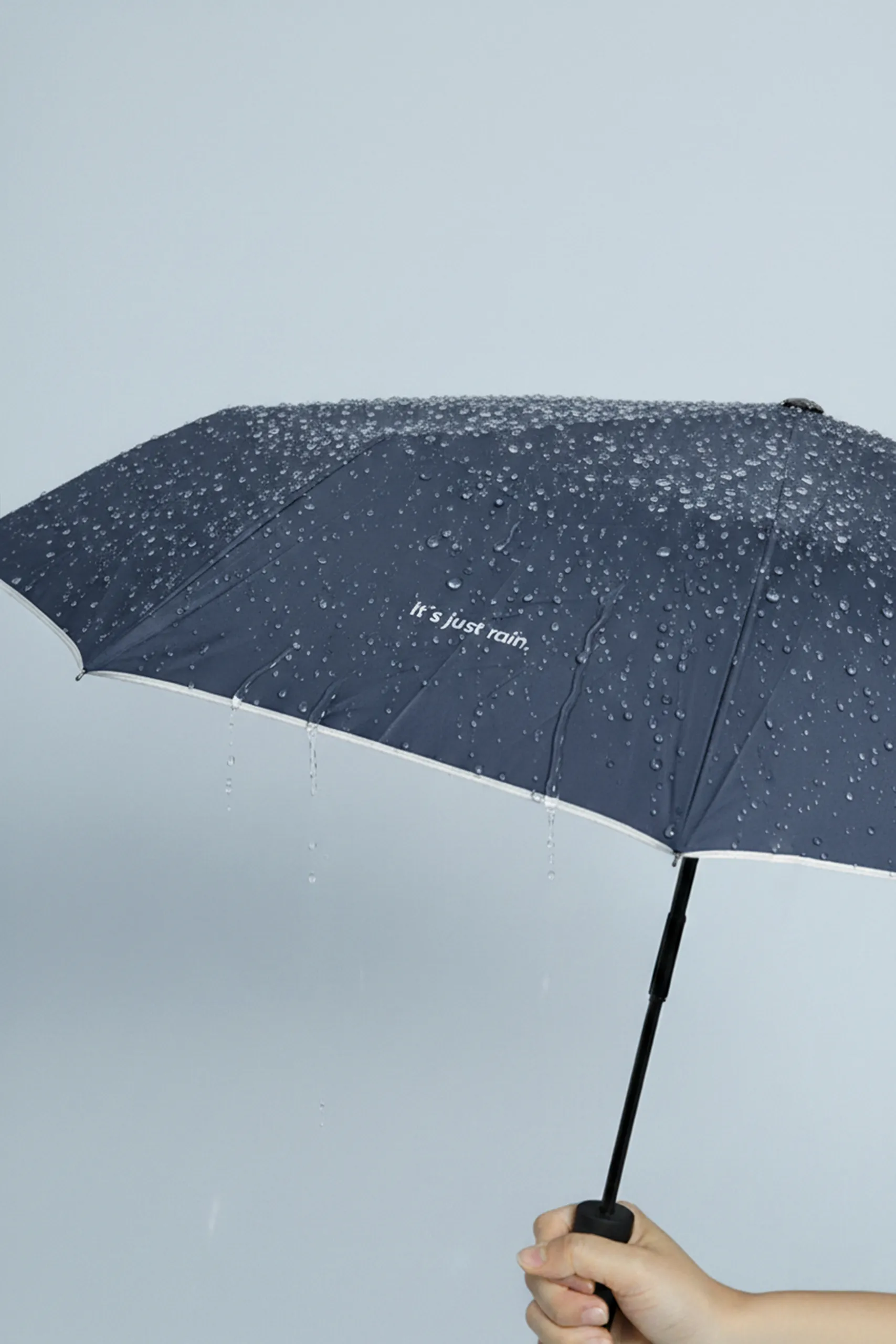 Paraguas azul oscuro con logo y linea reflectante. Con apertura automática y gotas por encima. Sujeto por una persona.